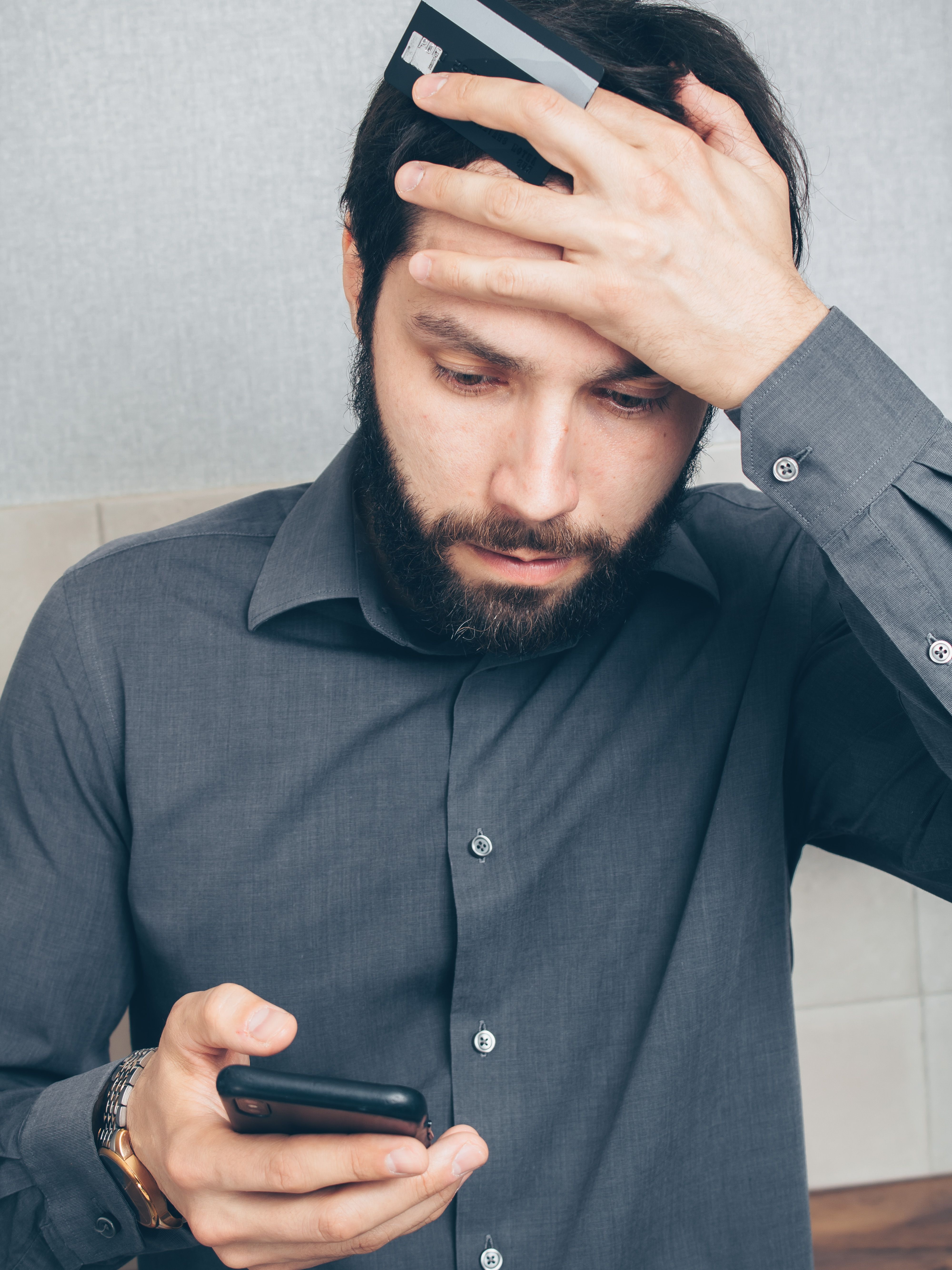 Man holds head and card in one hand and phone in other hand as he realises he has been a victim to bank impersonation fraud